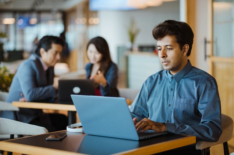Professional Using Laptop in Contemporary Shared Workspace with Team Meeting in Background.
