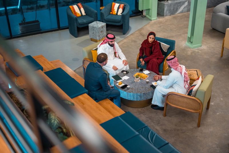 High-angle view of multicultural business meeting featuring young Middle Eastern men in traditional dress, Western businessman, and woman in burgundy hijab discussing in industrial-style lounge area.