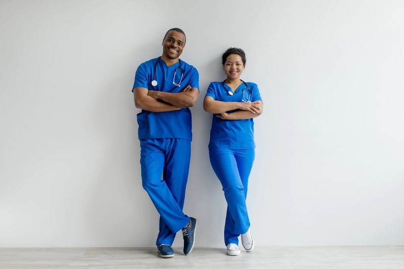 Portrait of two African American healthcare professionals in blue scrubs, confidently standing with arms crossed and smiling, posing together against a clean white studio background