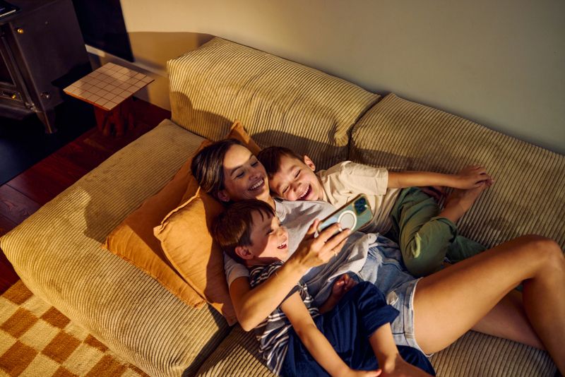 Happy mother and two sons lying on the sofa at home, watching smartphone and having fun