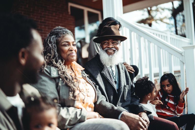 Affectionate and vibrant Black senior couple smile with gratitude while visiting with their multi-generation family on the front steps of their adult child's home.