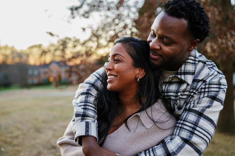 A beautiful woman of Indian descent and her Black husband affectionately embrace and smile while spending time outside at sunset.