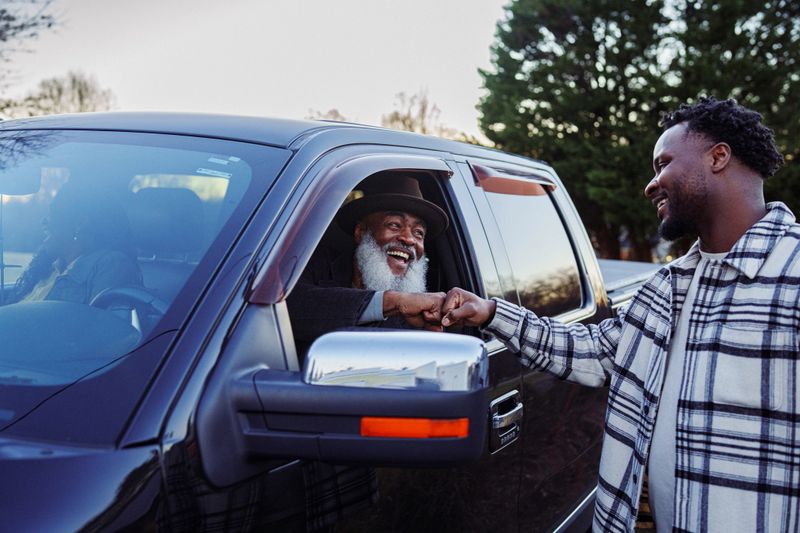 A joyful and active senior Black man sits in the driver's seat of a pickup truck and greets his adult son with a fist bump through the open window, after arriving at his son's house for a weekend family gathering.