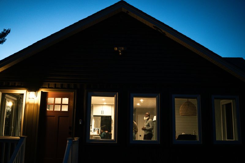 View shot from outside at night looking in the illuminated windows of a home at a Black senior man standing in the kitchen and talking with adult family members.