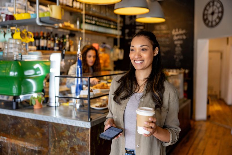 Happy woman leaving a cafe smiling after buying a cup of coffee to go and holding her cell phone - lifestyle concepts
