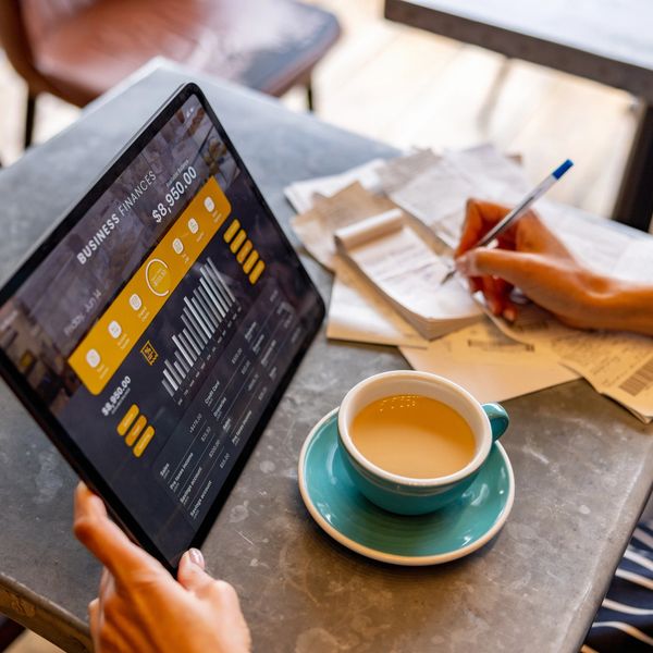 Person managing business finances on a tablet while making notes with a cup of coffee.