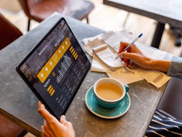 Person managing business finances on a tablet while making notes with a cup of coffee.