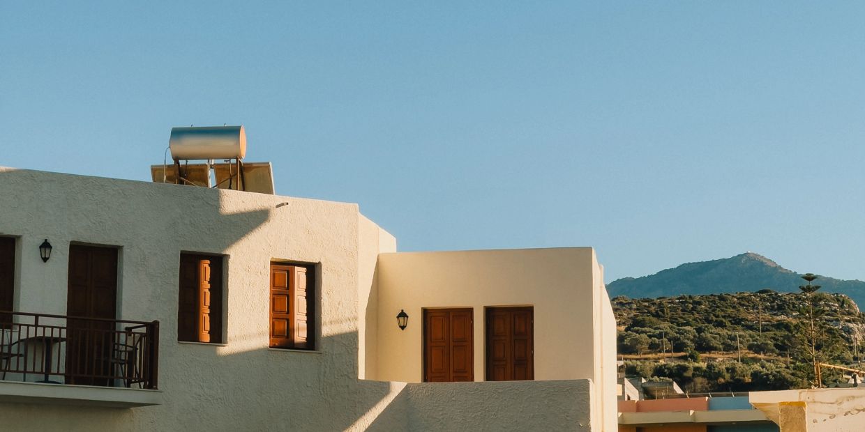 Sunlit Mediterranean-style building with wooden windows and mountain backdrop.
