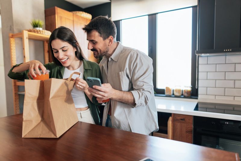 Young couple smiling and unpacking groceries from a paper bag in a modern kitchen, checking their food order on a smartphone while enjoying each other's company
