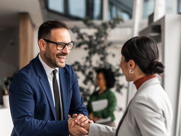 Two professionals shaking hands and smiling in an office setting.