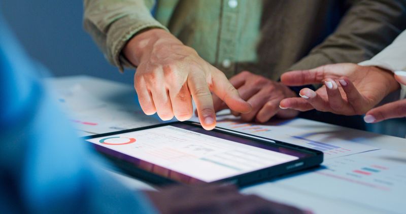 Close-up group of business working late, analyzing financial reports and charts on digital tablet surrounded by documents and graphs during strategic meeting at office night. Finance meeting concept.