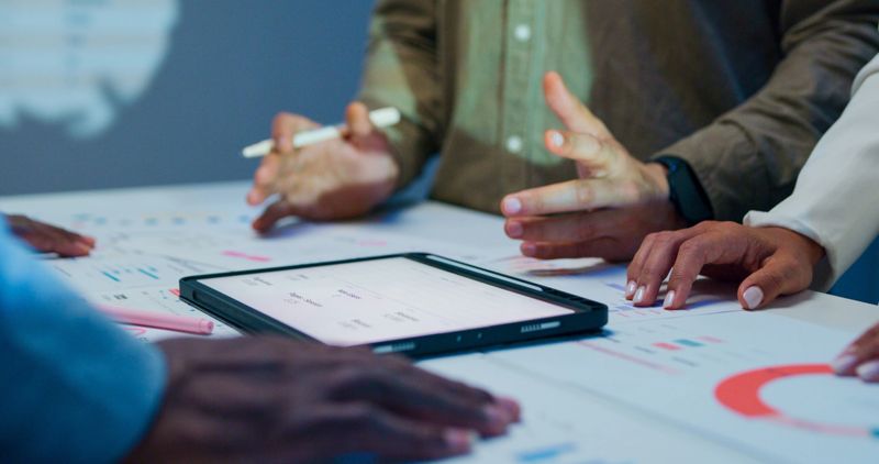 Close-up group of business working late, analyzing financial reports and charts on digital tablet surrounded by documents and graphs during strategic meeting at office night. Finance meeting concept.