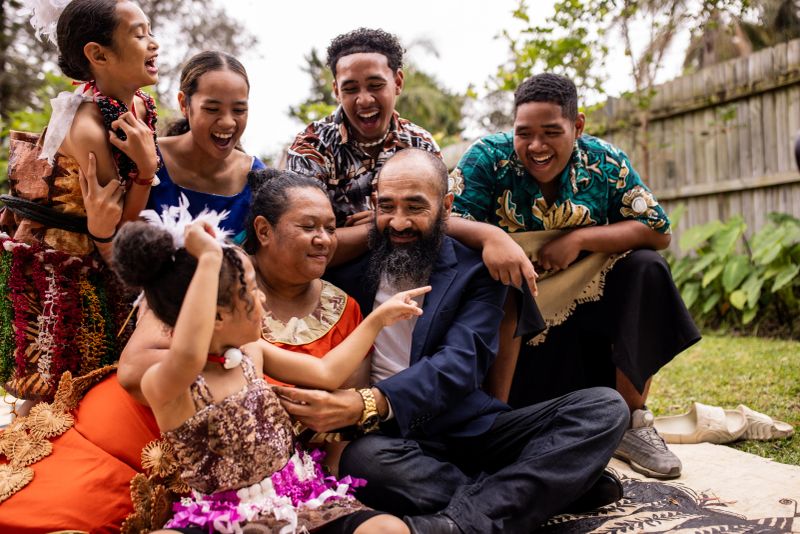 Large Pacific Islander family, spanning several generations, sits together on a carpet in their backyard, all wearing traditional clothing.