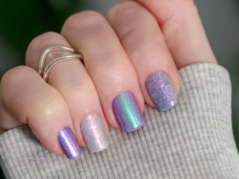 Skittles nails on hand. Colorful shiny manicure with different textures in light purple tones, pink and lilac hue. Closu-up of female hand with multicolored nails on green foliage background. Copy space