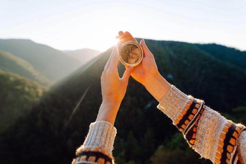 Rear view of young female traveler holding compass standing on cliff. Woman with hiking backpack and compass feeling freedom and enjoying mountain view. Travel and navigation concept.