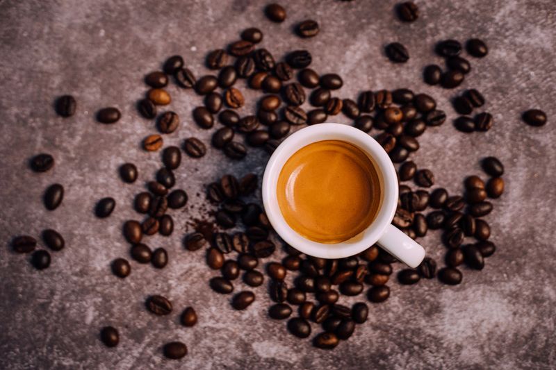 A beautifully composed overhead shot of a freshly brewed espresso in a white cup, set against a rustic, textured background. Surrounding the cup are scattered roasted coffee beans, adding depth and richness to the image.