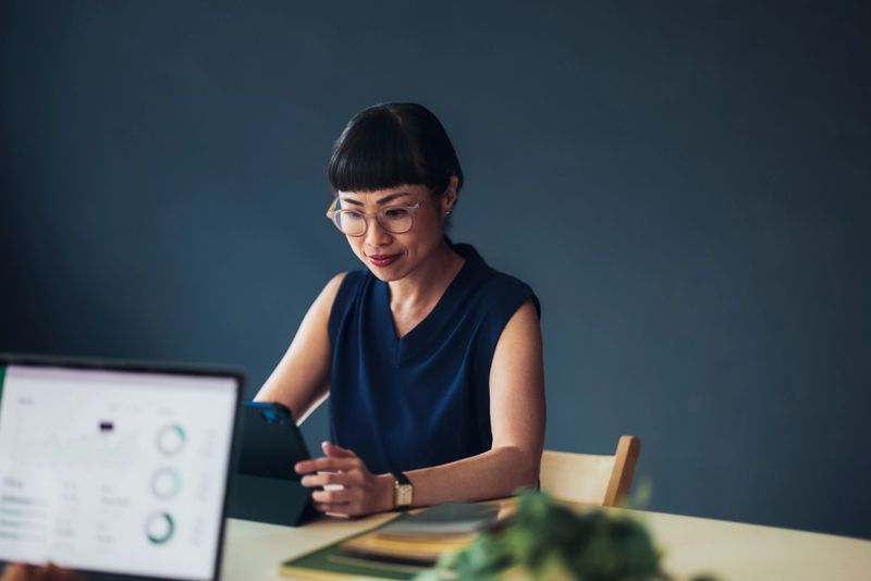 Focused woman using a tablet to work in a contemporary office, with charts visible on a laptop screen. Highlights themes of technology, productivity, and the modern workplace environment.