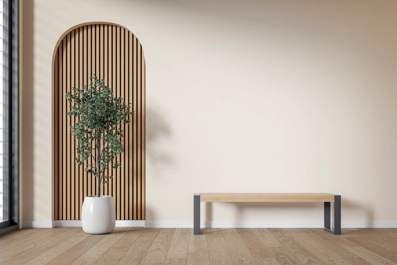 This photo shows a minimalistic interior space featuring a modern wooden bench with metal legs placed against a light beige (cream) wall. To the left, there's a tall green plant in a white pot positioned in front of an arched wooden slat wall accent, adding texture and visual interest. The flooring is a warm-toned wood, complementing the natural and calm aesthetic of the room. 3D rendered image.
