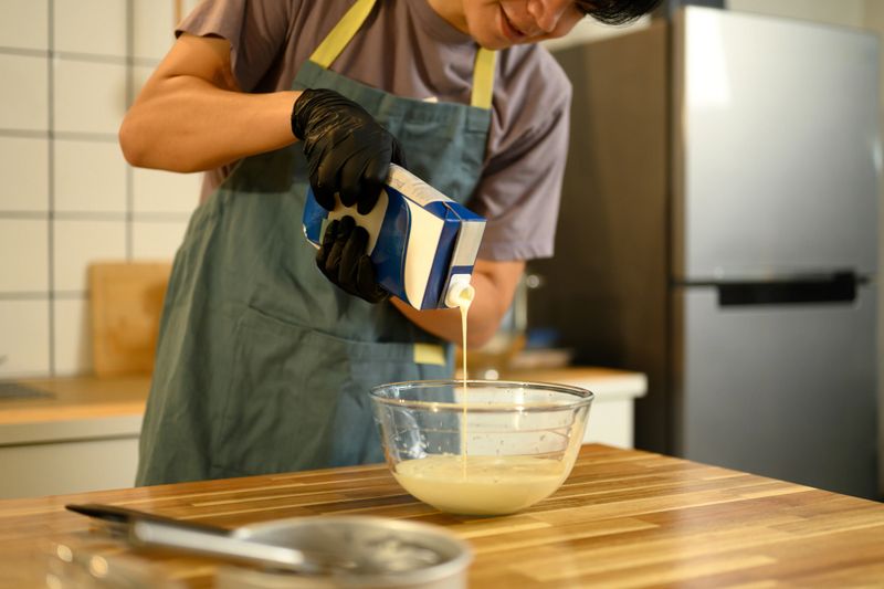 Man in apron and gloves pouring heavy cream into a glass bowl in preparation for making butter.