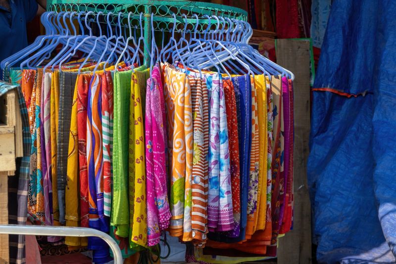 Colorful women's textile cotton sarees, including printed and batik designs, hanging on hangers at a shop front for tourist sale at the international trade fair in Kishoreganj.