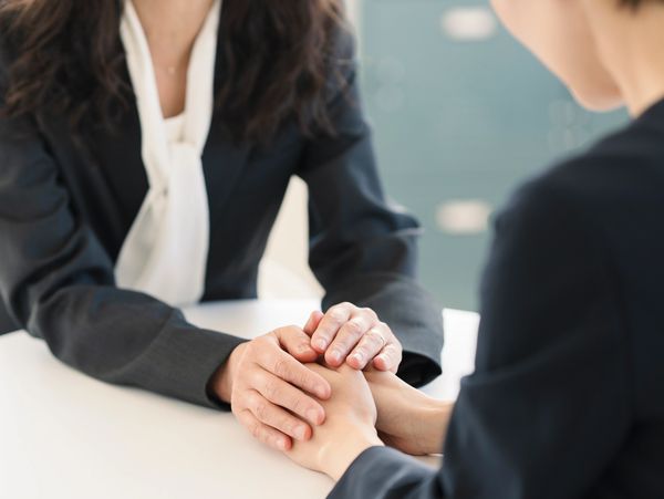 Two bookkeepers in business attire holding hands in a comforting gesture at a table.