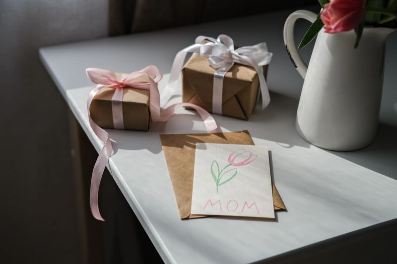 Handmade card with tulip drawing and MOM written on it rests on brown envelope, next to wrapped gifts and vase with pink tulips on white table. Concept of Mother's Day gifts, handmade with love