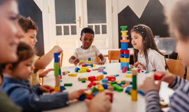 Diverse preschool children play with colorful building blocks at a classroom table, guided by a smiling teacher in a creative, inclusive early learning environment.