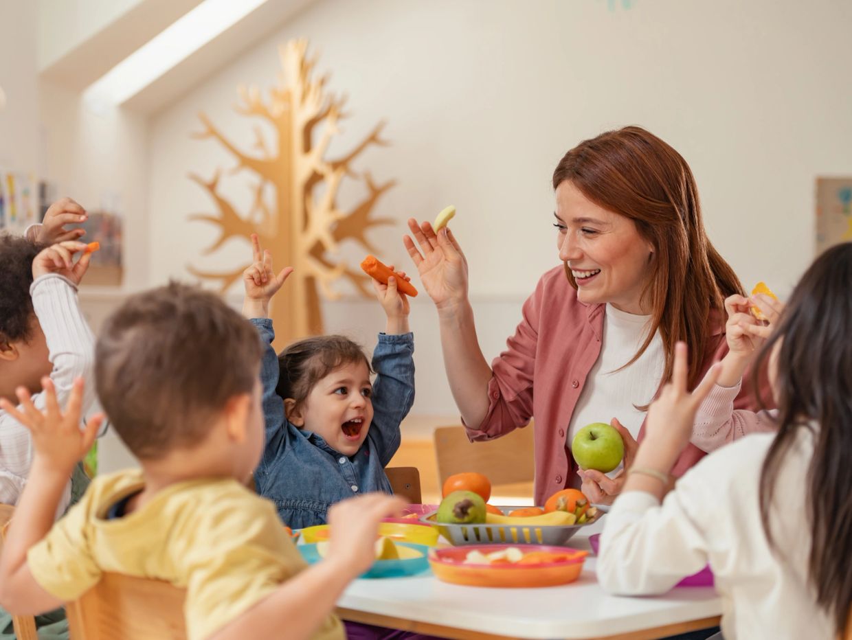 Teacher and children joyfully sharing healthy snacks around a table.