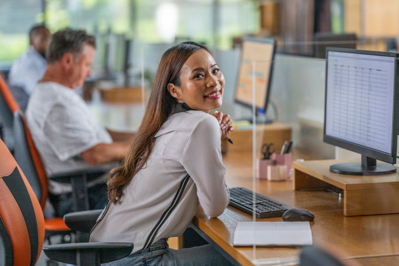 Smiling mid adult Asian businesswoman in a modern office, seated at her desk with a computer and organized documents, exuding positivity and professionalism.