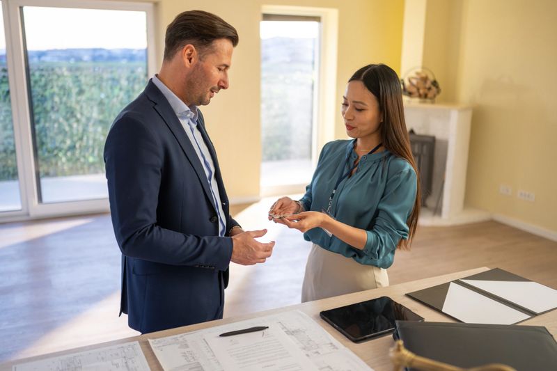 Professional portrait of mid-adult Asian female real estate agent wearing teal blouse exchanging documents with Caucasian male client in navy business suit inside sunlit contemporary home interior.