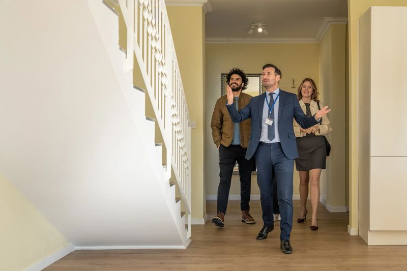 Mid adult Caucasian male real estate agent in a suit showing a modern home interior to a young diverse family, highlighting features with enthusiasm. The family includes a man and a woman appreciating the bright, spacious layout.