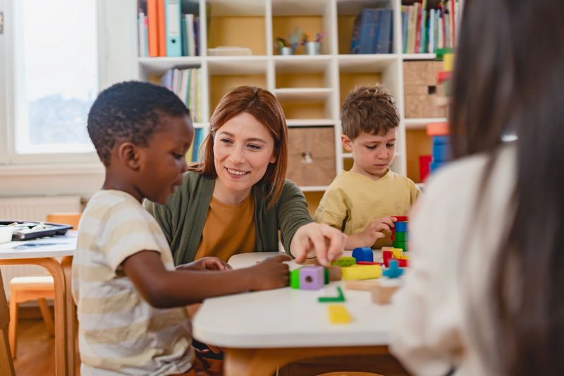 Preschool teacher interacts with a diverse group of children building with colorful blocks in a bright classroom, promoting creativity, teamwork, and multicultural early education.