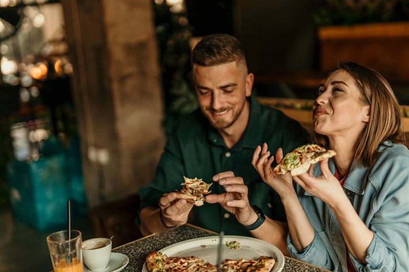 Happy young couple eating pizza slices at a restaurant table, enjoying their meal and each other's company