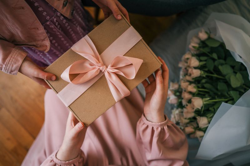 Close-up of a child giving a woman a gift box with a pink ribbon and a bouquet of delicate roses in soft natural light.