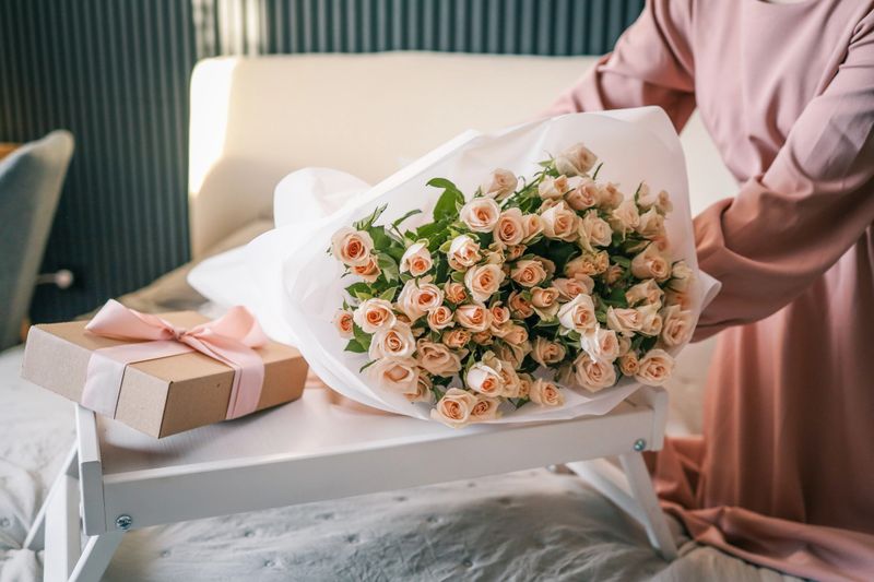 A wrapped gift with pink ribbon being handed over near a bouquet of roses and a cup of coffee on a bed, symbolizing care and appreciation.