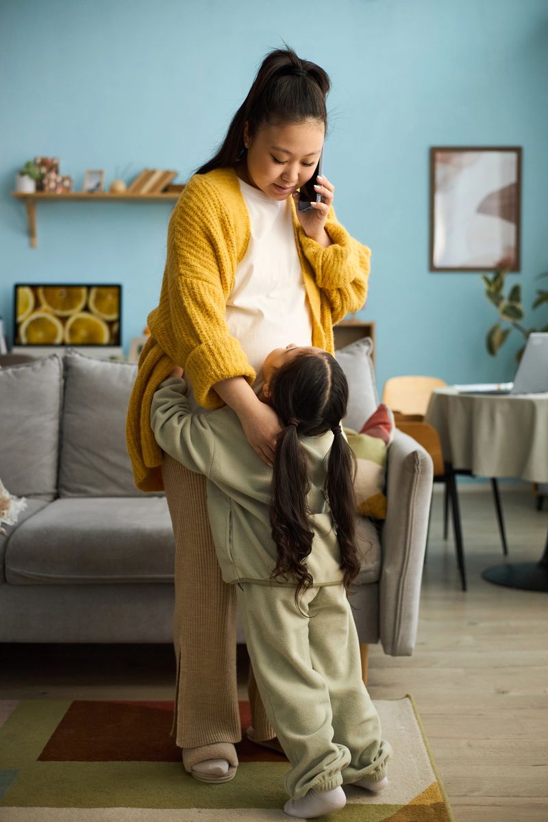 Asian mother multitasking by talking on phone while embracing her child in living room, surrounded by cozy decor and embracing furniture