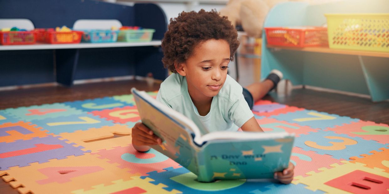 A child reading a book on a colorful alphabet mat.
