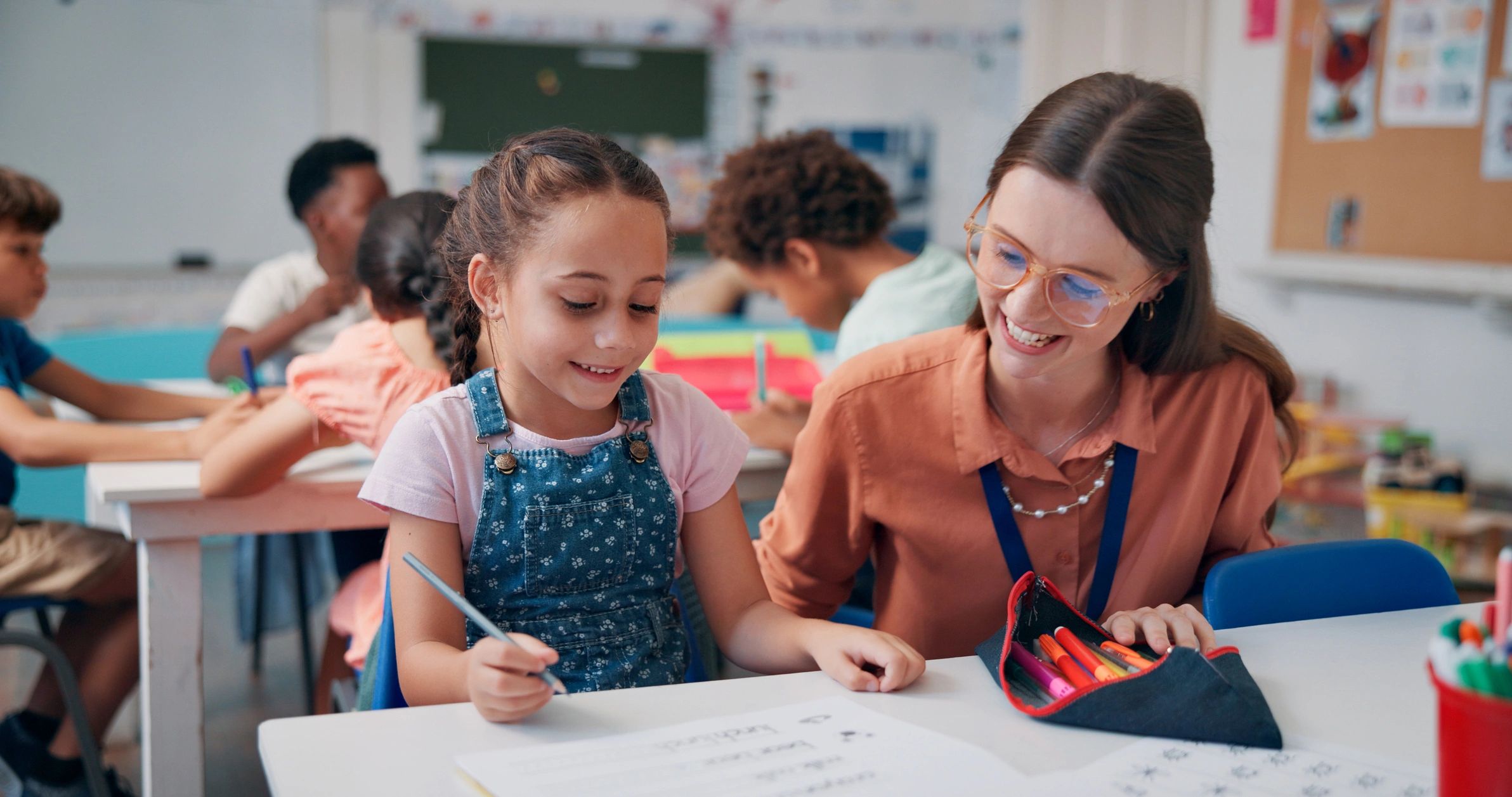 Teacher helping a smiling girl with her schoolwork in a classroom.