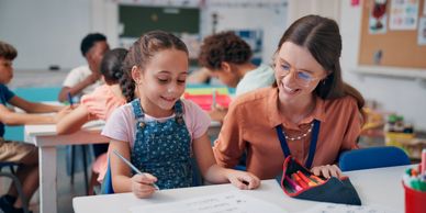 A teacher helps a young girl with her schoolwork in a classroom.