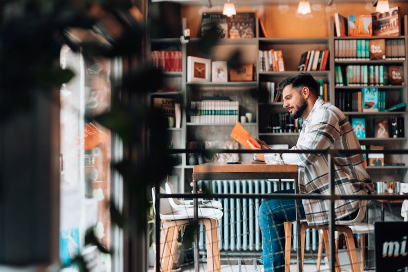 Young bearded man sitting at a table in a bookstore cafe, savoring the pleasure of reading a book while surrounded by a warm, inviting atmosphere filled with knowledge and inspiration