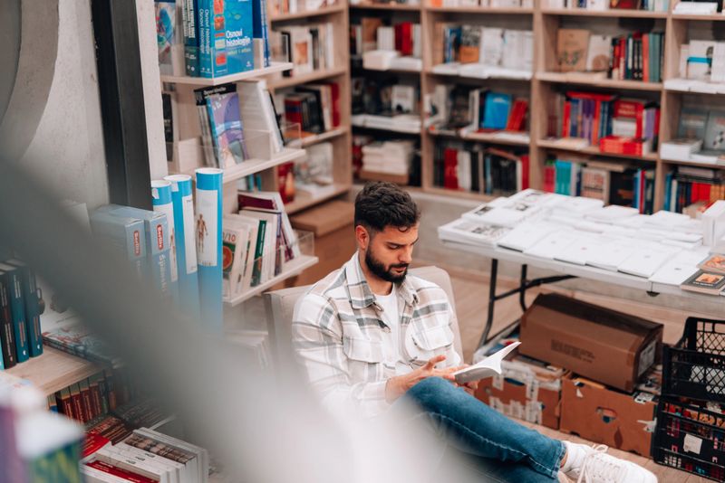 Young bearded man sitting on a gray armchair, enjoying reading a book in a cozy bookstore full of books, enjoying the quiet atmosphere and the pleasure of reading