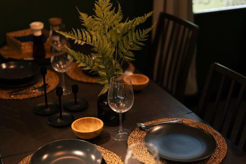 Elegant table is set with black plates, wooden bowls, wine glasses, candles, and a centerpiece of ferns, ready for a sophisticated dinner party
