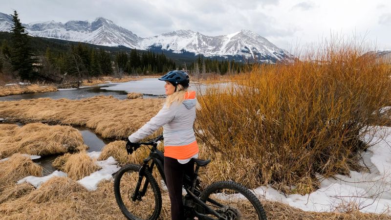 Mature woman with e-bike pauses in mountain meadow near creek in winter, looks towards snowcapped mountains at end of valley, Crowsnest Pass, Alberta
