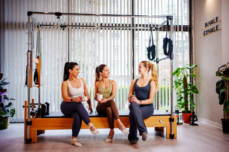 Three women standing and chatting near Pilates equipment in a sunlit fitness studio. The group includes a Pilates instructor and two students, all dressed in activewear, enjoying a casual conversation after a workout session. The modern studio setting, with floor-to-ceiling windows and indoor plants, creates a fresh and motivational atmosphere. This image reflects wellness, community, and a healthy lifestyle.