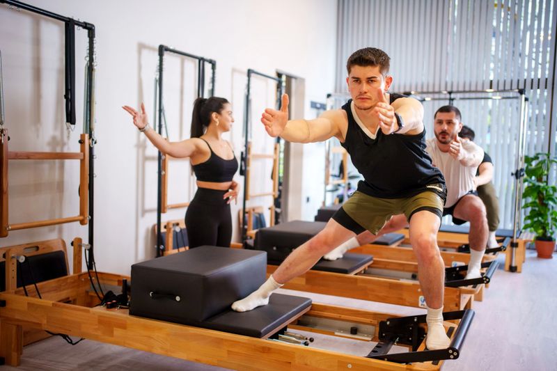 Pilates Instructors working at Pilates Saloon. Young men practicing Pilates system on reformer supervised by qualified trainer