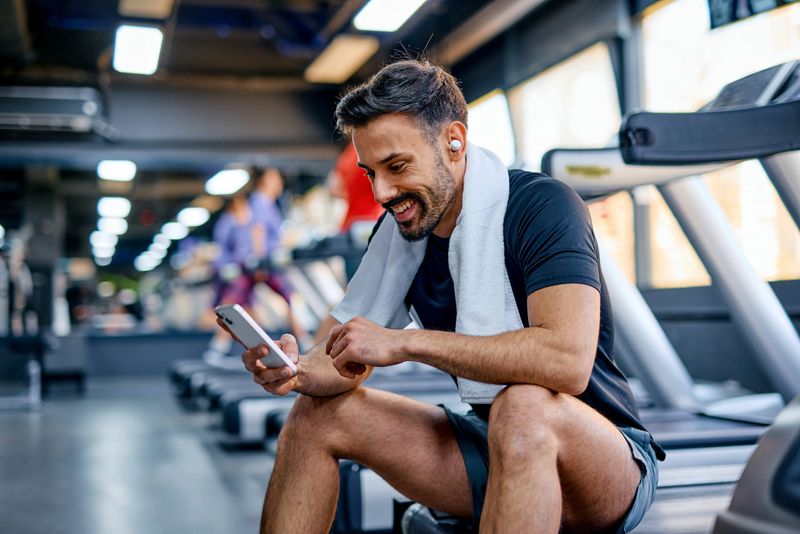 Mid adult sportsman is sitting on a treadmill in a gym, wearing earbuds and a towel around his neck, looking at his smartphone and smiling after his workout