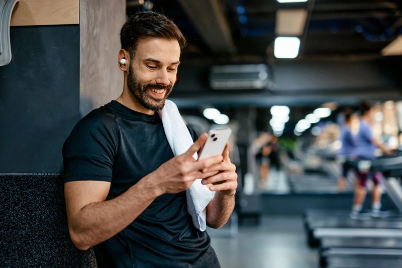 Happy athlete wearing wireless earbuds, engaging with a smartphone at the gym, checking messages, switching music, or utilizing a fitness app while enjoying a workout