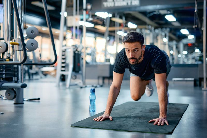 Young sportsman wearing earphones is performing mountain climbers on a fitness mat during an intense cross training workout in a modern gym, showcasing determination and strength