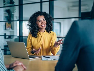 A woman in a yellow blouse smiling and talking during a meeting.