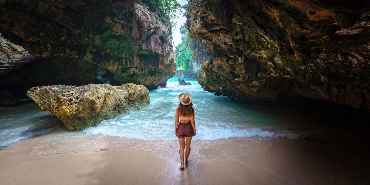 A woman stands at the entrance of a rocky cave by the beach.